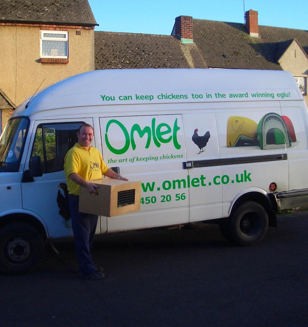 Ein lieferfahrer neben einem lieferwagen der marke Omlet, der eine kiste mit hühnern transportiert